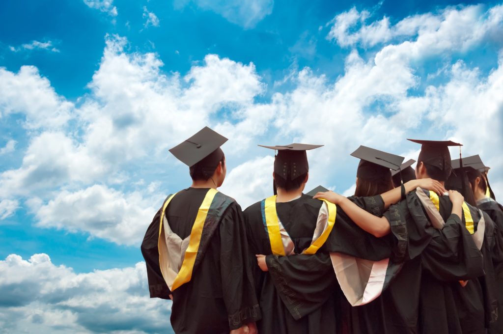 Back of four graduates, looking at blue/cloudy sky with grad caps and gowns on
